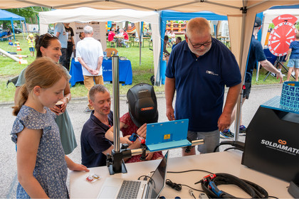 Vier Personen stehen vor einem virtuellen Schweißgerät unter einem Pavillon und beobachten dabei wie ein Kind schweißen übt. Auf dem Tisch liegen elektronische Bauteile, Kabel und ein Laptop, im Hintergrund sind weitere Besucher und Marktstände zu sehen.