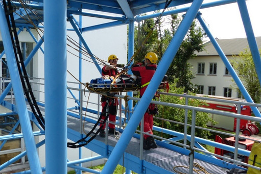 Zwei Personen in Schutzkleidung und gelben Helmen sichern eine Trage mit Seilen in einem blauen Stahlgerüst, im Hintergrund sind Gebäude und Bäume sichtbar.