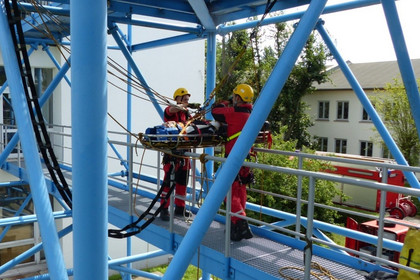 Zwei Personen in Schutzkleidung und gelben Helmen sichern eine Trage mit Seilen in einem blauen Stahlgerüst, im Hintergrund sind Gebäude und Bäume sichtbar.
