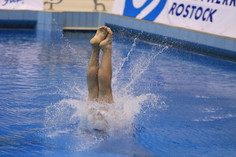 Ein Turmspringer taucht senkrecht mit gestreckten Beinen ins Wasser ein, während eine große Spritzfontäne den Moment des Eintauchens begleitet.