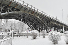 Eine große Brücke aus Altstahl überspannt eine schneebedeckte Landschaft. Der Boden ist vollständig mit Schnee bedeckt, vereinzelt stehen kleine Büsche oder Bäume im Vordergrund.