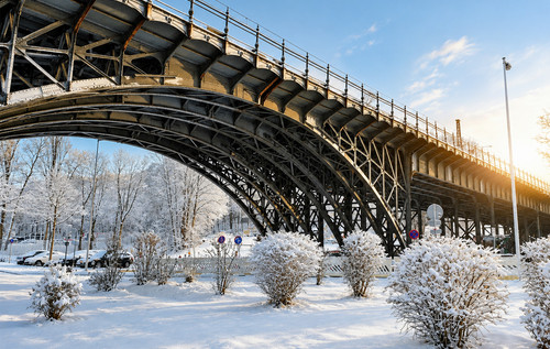 Eine große Brücke aus Altstahl überspannt eine schneebedeckte Landschaft. Der Boden ist vollständig mit Schnee bedeckt, vereinzelt stehen kleine Büsche oder Bäume im Vordergrund.