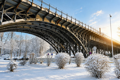 Eine große Brücke aus Altstahl überspannt eine schneebedeckte Landschaft. Der Boden ist vollständig mit Schnee bedeckt, vereinzelt stehen kleine Büsche oder Bäume im Vordergrund.