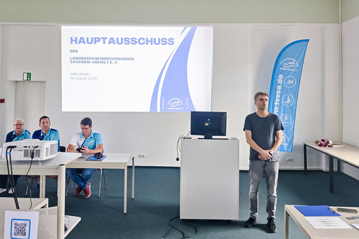Ein Mann steht neben einem Rednerpult vor einer Präsentationsfolie mit der Aufschrift „Hauptausschuss des Landesschwimmverbandes Sachsen-Anhalt e. V.“; drei Personen in blauen Vereinsshirts sitzen am Tisch, daneben ein blauer Roll-Up.