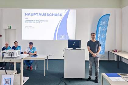 Ein Mann steht neben einem Rednerpult vor einer Präsentationsfolie mit der Aufschrift „Hauptausschuss des Landesschwimmverbandes Sachsen-Anhalt e. V.“; drei Personen in blauen Vereinsshirts sitzen am Tisch, daneben ein blauer Roll-Up.