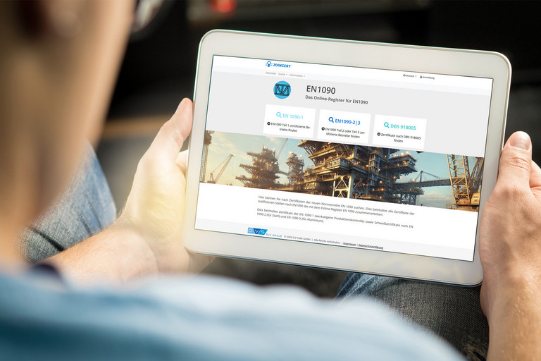 Person hält ein Tablet in den Händen und betrachtet darauf eine Webseite mit Bildern und Texten, der Bildschirm ist deutlich sichtbar und zeigt die Norm EN 1090, der Hintergrund unscharf.