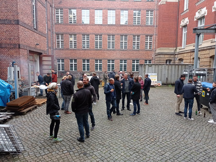Gruppe von etwa 30 Personen steht in einem Innenhof mit Kopfsteinpflaster zwischen roten Backsteingebäuden, einige unterhalten sich, andere stehen an einem Grillplatz