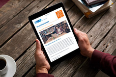 Eine Person hält ein Tablet in den Händen, auf dem ein Newsletter der DVS SLV Halle mit dem Titel „Bildungskurier“ angezeigt wird; das Gerät liegt auf einem rustikalen Holztisch neben einer Tasse Kaffee.