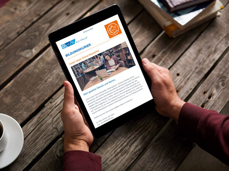 Eine Person hält ein Tablet in den Händen, auf dem ein Newsletter der DVS SLV Halle mit dem Titel „Bildungskurier“ angezeigt wird; das Gerät liegt auf einem rustikalen Holztisch neben einer Tasse Kaffee.