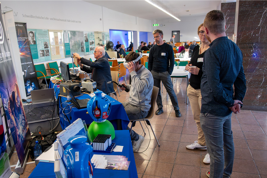Im Foyer der SLV Halle erleben mehrere Personen moderne Schweißtechnik mit Virtual-Reality-Brille und Demonstrationsgeräten. Ein älterer Mann justiert eine Versuchsanlage, während andere interessiert zusehen und sich austauschen. Im Hintergrund sind weitere Gäste an Tischen im Gespräch.