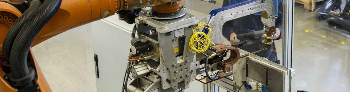 Ein großer Industrieroboter mit Schweißvorrichtung befindet sich in einer automatisierten Fertigungszelle, im Hintergrund steht ein Mann in Arbeitskleidung an einem Steuerpult hinter einer Glaswand.