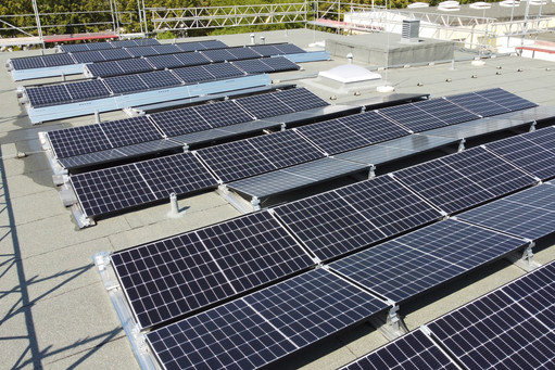 Mehrere Reihen von Solarpaneelen sind auf einem Flachdach installiert, umgeben von einem Sicherheitsgeländer; im Hintergrund befinden sich technische Aufbauten und Dachauslässe.