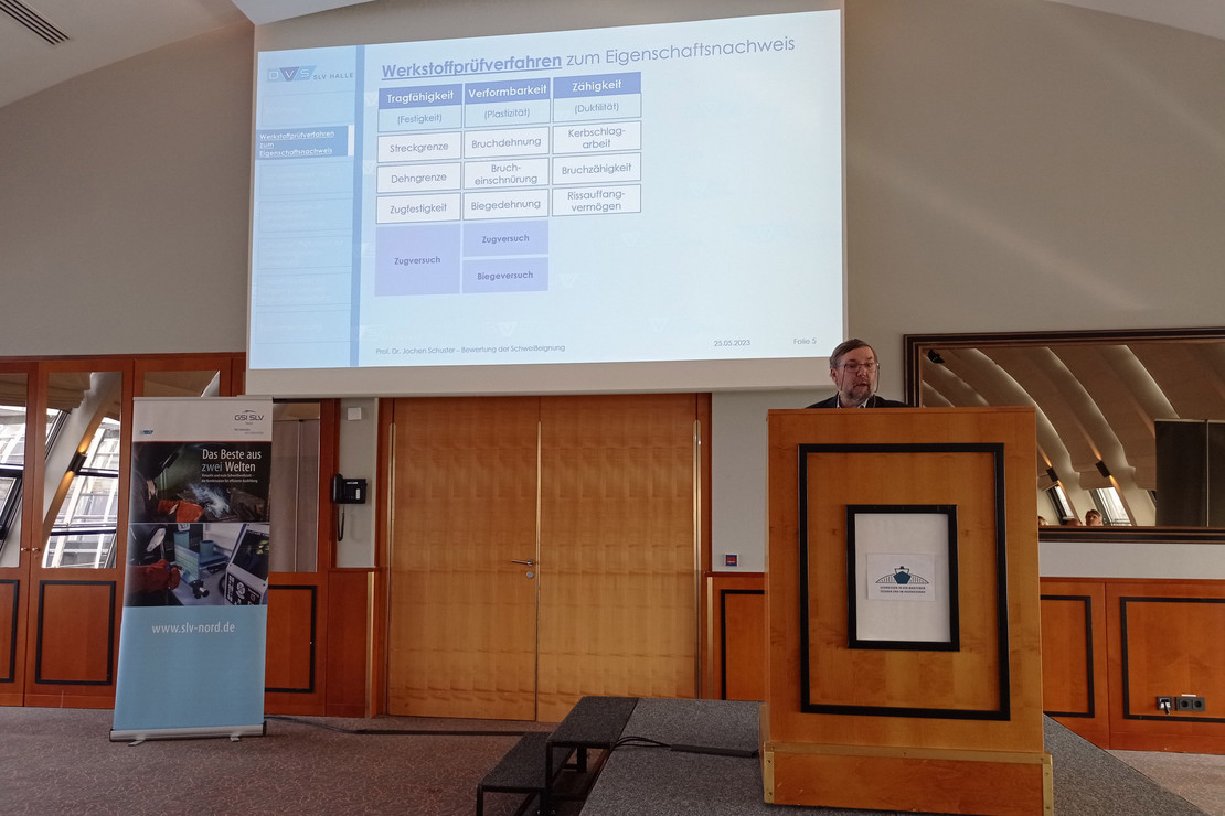 Mann steht an einem Rednerpult in einem Konferenzraum, hinter ihm eine Leinwand mit Präsentation zu Werkstoffprüfverfahren, links ein Roll-Up mit SLV-Halle-Motiv