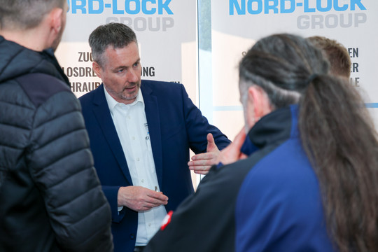 Mann im Gespräch mit zwei Personen an einem Messestand, er zeigt auf ein Exponat, im Hintergrund der Schriftzug NORD LOCK