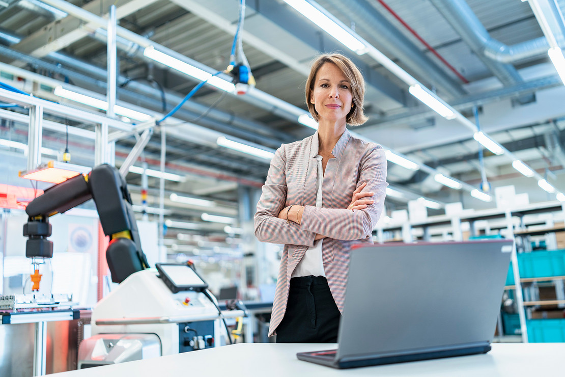 Frau mit überkreuzten Armen steht in einer Industriehalle mit Robotern vor einem Laptop.