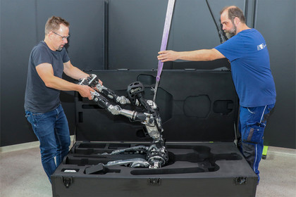Zwei Männer heben einen humanoiden Roboter mithilfe eines violetten Hebegurts aus einem geöffneten Transportkoffer. Einer hält die Arme des Roboters, während der andere den Hebegurt führt.