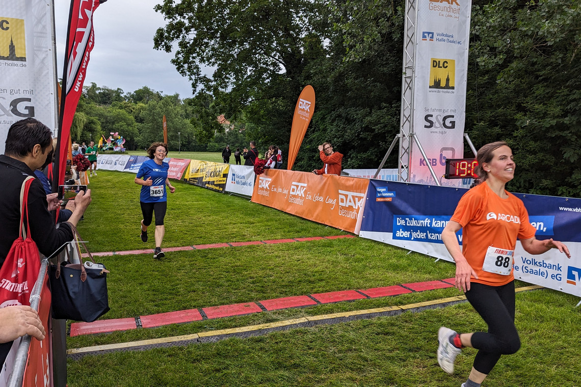 Zwei Frauen überqueren nacheinander die Ziellinie beim Firmenlauf in Halle – vorne in orange, dahinter in blau. Links stehen jubelnde Zuschauer\:innen und Fotograf\:innen hinter Absperrungen, rechts sind Sponsorenbanner und eine digitale Uhr mit der Zeit 1:00:34 sichtbar.