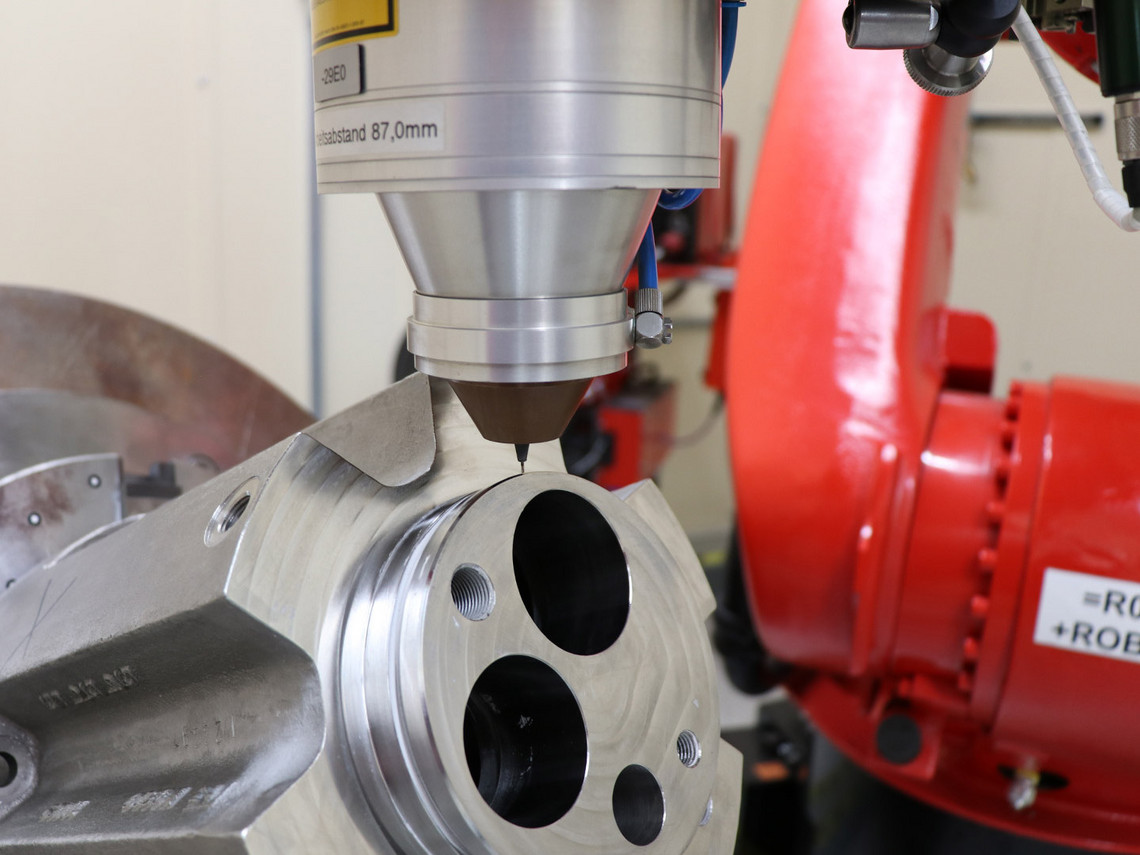 Nahaufnahme eines Metallbauteils mit Bohrungen, das unter einem Additivkopf bearbeitet wird. Der Düsenabstand zum Werkstück ist präzise eingestellt. Im Hintergrund ist der rote Roboterarm sichtbar.