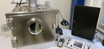 Elektronenstrahlbearbeitung: Ein kompaktes Vakuumkammer-System aus Edelstahl mit Sichtfenster steht auf einem Labortisch. Daneben befinden sich eine Steuerkonsole, eine Handfernbedienung und ein Monitor. Oben auf der Kammer sind Sensoren und Anschlüsse für Vakuumtechnik montiert.
