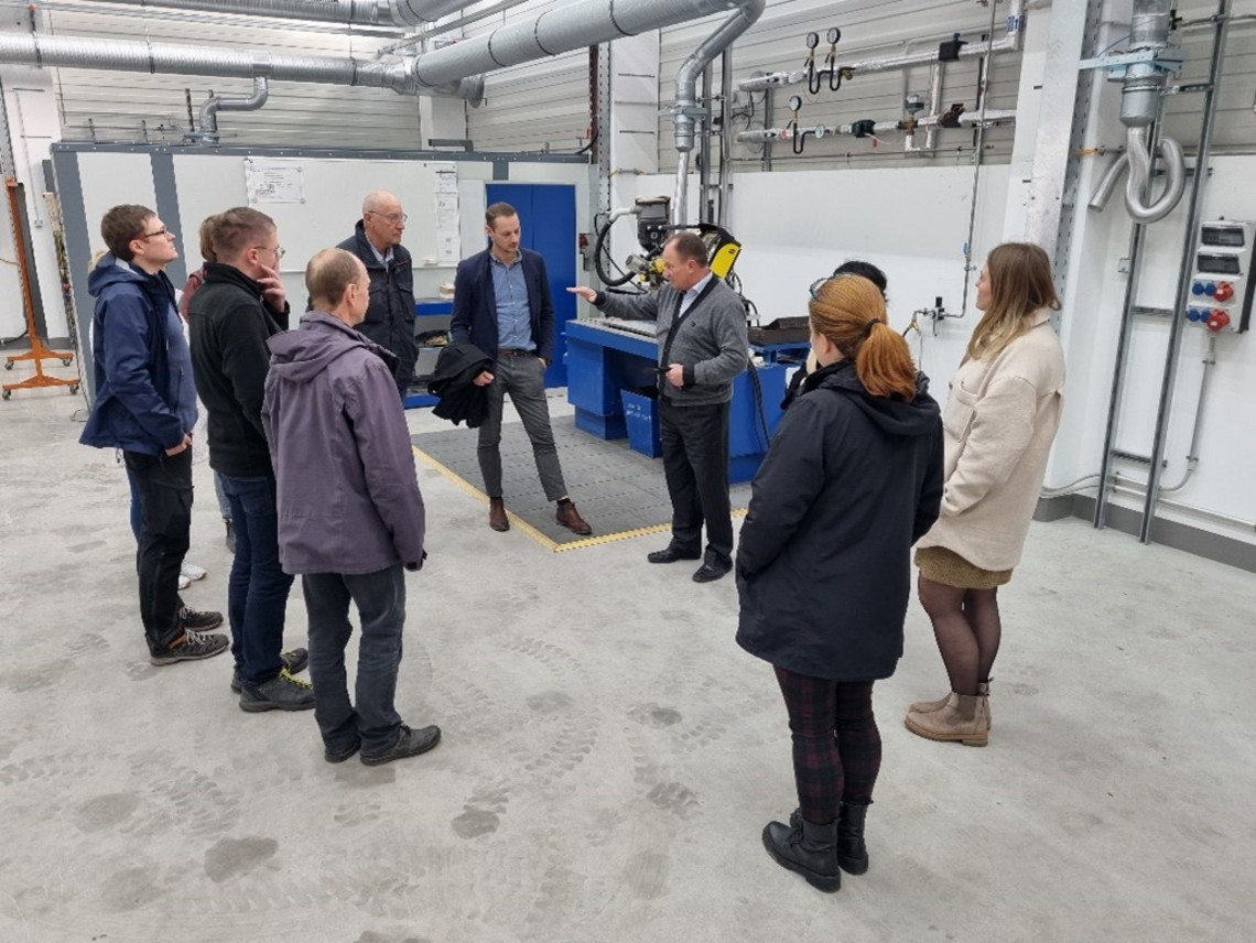 Zehn Personen, darunter drei Frauen und sieben Männer, stehen in einem modernen Technikraum im Halbkreis um einen Mann im Anzug, der eine Maschine erklärt; im Hintergrund sind Lüftungsrohre und Schweißtechnik sichtbar.