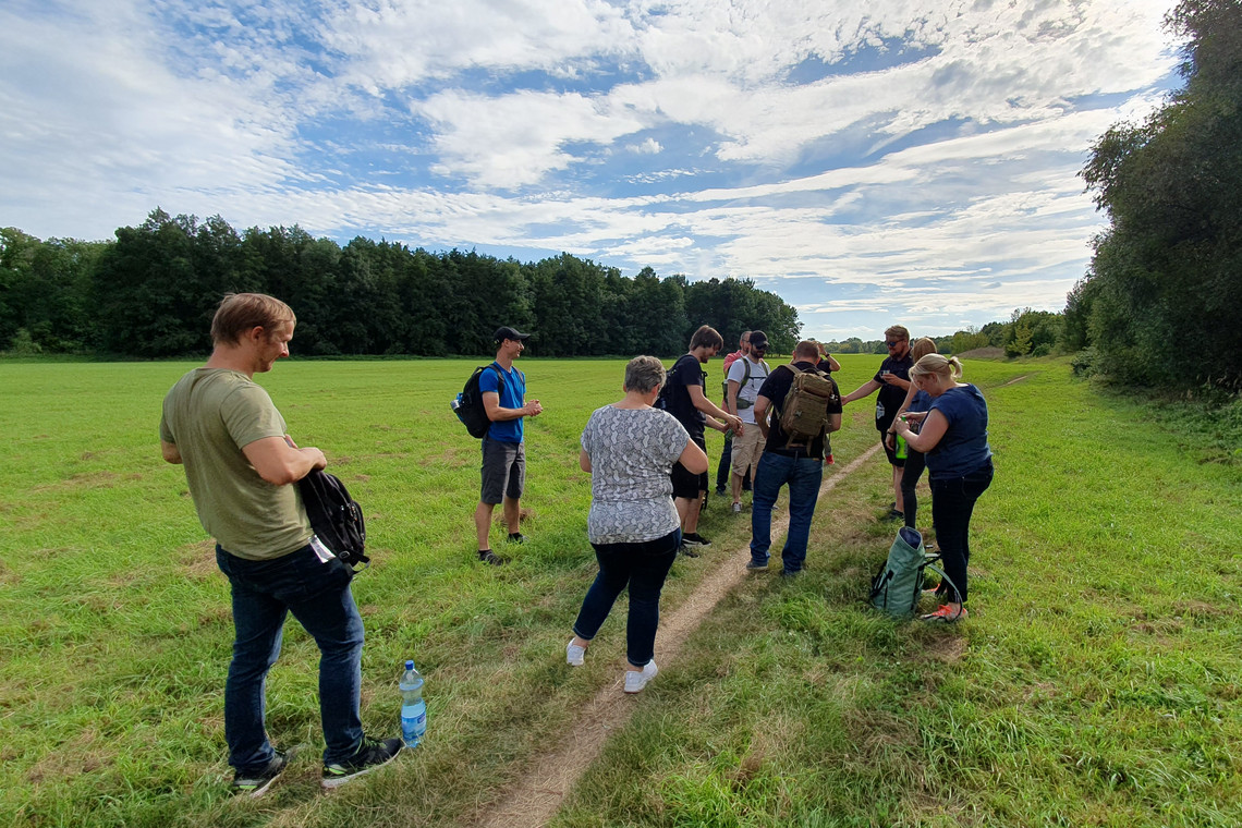 Gruppe von Wandernden steht auf einem schmalen Pfad inmitten einer weiten grünen Wiese, einige richten Rucksäcke oder greifen zur Wasserflasche, am Horizont beginnt ein dichter Wald unter teils bewölktem Himmel.
