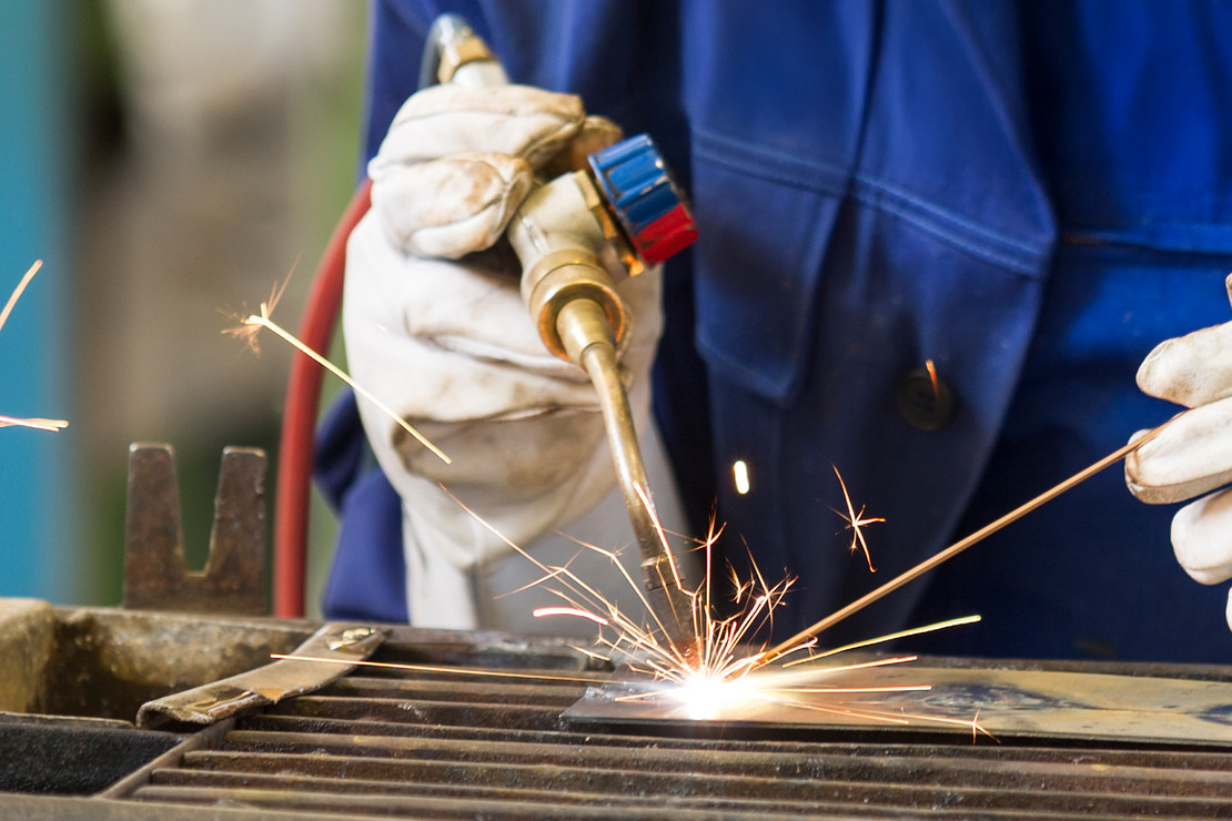 Hände in Schutzhandschuhen führen eine Schweißflamme auf ein Metallstück, Funken sprühen hell auf, im Hintergrund blaue Arbeitskleidung.