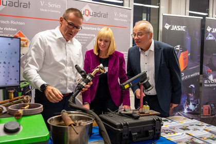 Drei Personen stehen an einem Messestand und betrachten gemeinsam ein technisches Exponat. Eine Frau in violettem Blazer hält ein Werkzeug in der Hand, zwei Männer beobachten interessiert. Im Hintergrund sind Firmenbanner und Geräte zu sehen.