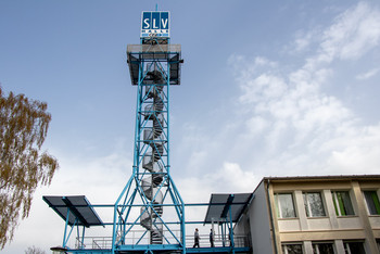 der blaue SLV Turm ragt in die Höhe, drei Männer tragen eine Fensterscheibe vom Plateau in das verbundene Obergeschoss eines Gebäudes