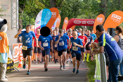 Gruppe von Läuferinnen und Läufern beim Start eines Firmenlaufs, vorn laufen fünf Personen (vier Männer, eine Frau) in blauen Shirts, Zuschauer stehen rechts, links steht ein Maskottchen in orangefarbener Kleidung.