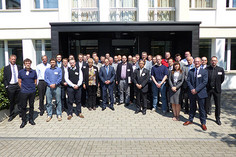 Eine große Gruppe steht bei sonnigem Wetter für ein Gruppenfoto vor einem Institutsgebäude mit heller Fassade.