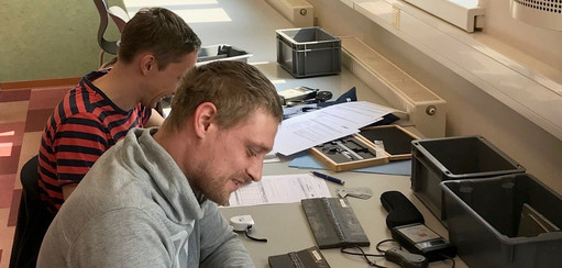 Zwei Männer sitzen nebeneinander an einem Tisch in einem hellen Schulungsraum und bearbeiten konzentriert Unterlagen mit Lineal und Taschenrechner. Auf dem Tisch liegen weitere Ordner und Prüfmittel.