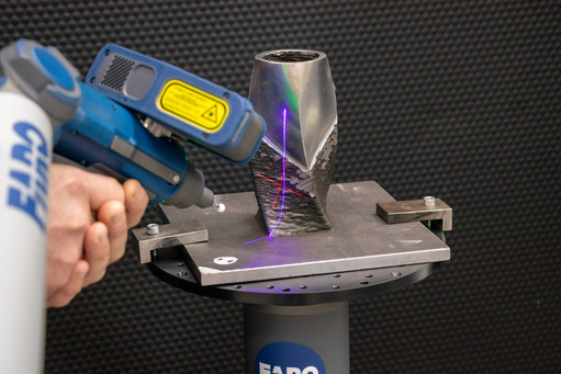 Ein strukturierter Metallkörper mit unregelmäßiger Oberfläche wird mit einem tragbaren 3D-Laserscanner vermessen; das Bauteil ist auf einem FARO-Messtisch fixiert, während der Scanner farbige Laserlinien projiziert.