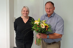 Eine Frau mit weißblondem Haar und ein Mann mit Kurzarmhemd stehen nebeneinander vor einer weißen Wand; der Mann hält einen bunten Blumenstrauß in einer Glasvase. Beide lächeln.