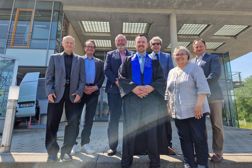 Sieben Personen stehen bei Sonnenschein auf einer Treppe vor einem modernen Institutsgebäude; in der Mitte trägt ein Mann einen Talar mit blauer Schärpe und lächelt in die Kamera.