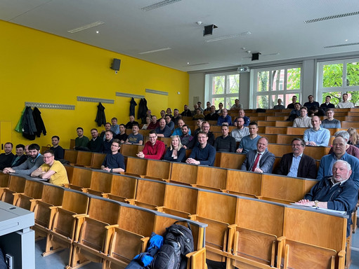 Ein alter Hörsaal mit Holzbänken und Klapptischen. Die Sitze sind ca. ein Drittel belegt von erwachsenen Personen und Freizeit- und Businesskleidung.