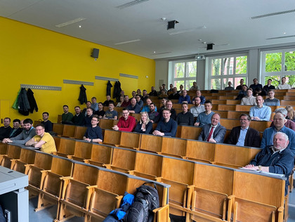 Ein alter Hörsaal mit Holzbänken und Klapptischen. Die Sitze sind ca. ein Drittel belegt von erwachsenen Personen und Freizeit- und Businesskleidung.