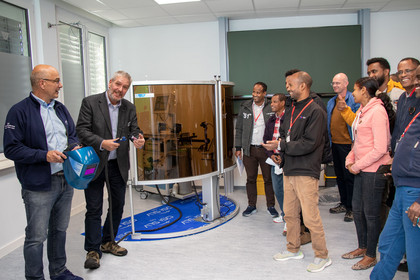 Eine Gruppe steht um ein technisches Gerät mit Schutzschild in einem Schulungsraum, ein Mann erklärt etwas und hält einen Schweißhelm, die Zuhörenden lächeln und hören aufmerksam zu.