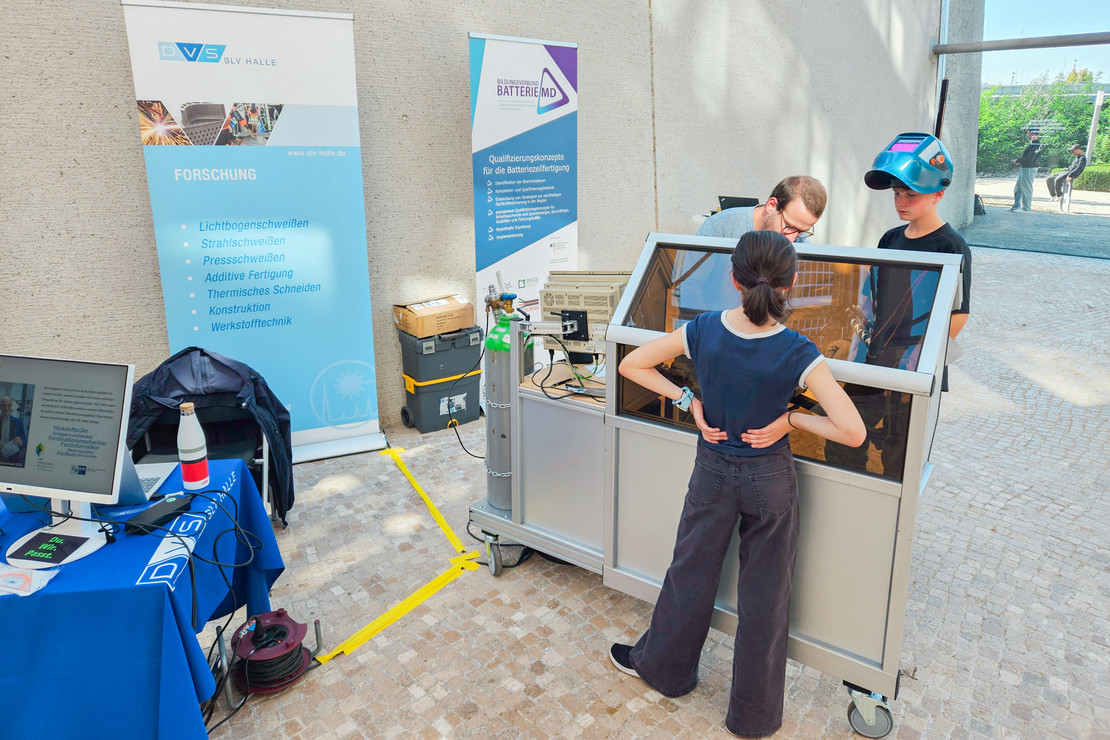 Ein Messe- oder Infostand der SLV Halle und des Projekts BatterieMD mit Bannern, Computer und Versuchsaufbau, an dem junge Personen mit Schweißhelm und Begleitung ein Verfahren ausprobieren.