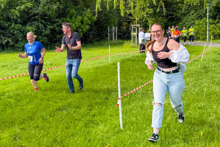 Eine Frau läuft neben einer abgesperrten Strecke über eine Wiese in einem Park. Auf der Strecke selbst laufen eine Frau mit Startnummer und ein weiterer Mann, der applaudierend neben ihr läuft. Alle sind glücklich. Im Hintergrund stehen mehrere Personen am Wegesrand vor einer Baumreihe.