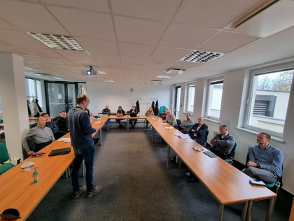 Ein Mann steht in einem U-förmig bestuhlten Seminarraum vor einer Gruppe aus neun Männern und drei Frauen, die ihm aufmerksam zuhören; auf den Tischen liegen Unterlagen und Getränke.