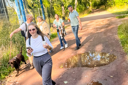 Vier Personen gehen auf einem unbefestigten Weg zwischen Bäumen, im Vordergrund hält eine Frau mit Sonnenbrille ein Smartphone in der Hand. Links läuft ein Hund am Wegesrand, auf dem Boden befindet sich eine größere Pfütze.