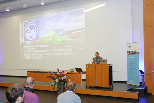 Vortragssaal mit Redner am Podium und einer Begrüßungsfolie auf der Leinwand, Thema: Weiterbildung im Schienenfahrzeugbau