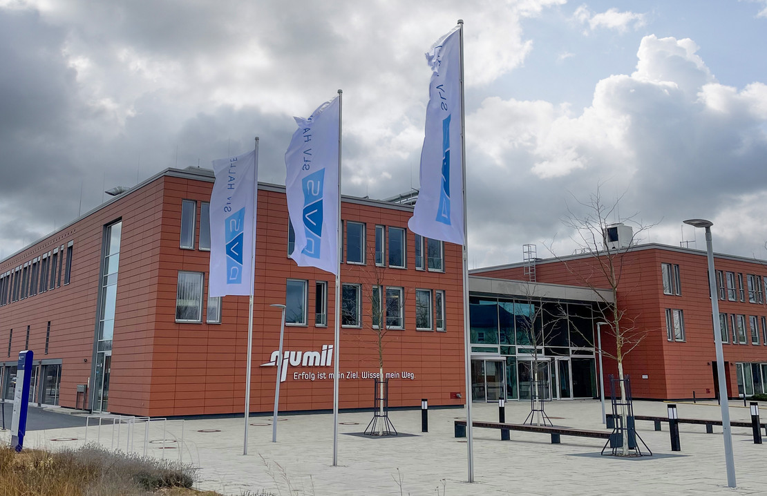 Modernes orangefarbenes Gebäude mit Glasfassade, davor drei wehende Flaggen mit SLV-Halle-Logo und Aufschrift „njumii – Das Bildungszentrum des Handwerks“, bewölkter Himmel im Hintergrund