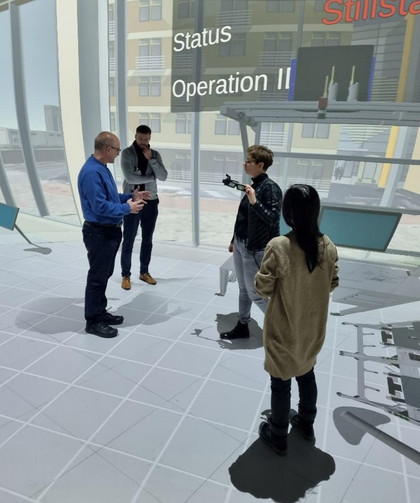 Vier Personen diskutieren in einem virtuellen Raum mit Boden- und Wandprojektionen über einen digitalen Industrieprozess; hinter ihnen sind Statusanzeigen mit Begriffen wie „Status“ und „Operation II“ zu sehen.