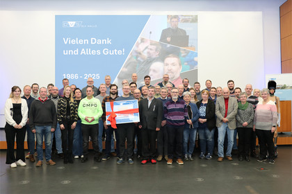 Eine größere Gruppe von Personen steht nebeneinander in einem Veranstaltungsraum vor einer beleuchteten Bühne mit Leinwand und Präsentationsgrafik im Hintergrund.