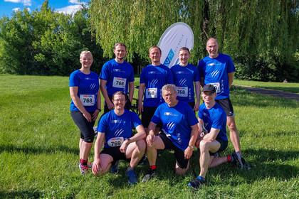 Sieben Männer und eine Frau in blauen T Shirts posieren gemeinsam auf einer Wiese im Freien, hinter ihnen eine Beachflag von der SLV Halle GmbH. Einige stehen, die anderen knien davor, im Hintergrund sind Bäume und dichter Bewuchs zu sehen.