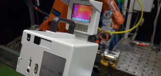 Ein Infrarot-Messgerät mit Bildschirm zeigt ein Wärmebild, im Hintergrund sind ein orangefarbener Industrieroboter und ein metallischer Prüftisch zu sehen.