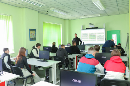 Klassenraum mit zehn jungen Erwachsenen, die an Computern sitzen, ein Dozent steht vorne an einem interaktiven Whiteboard mit Präsentationsfolie, grüne Wände und Jalousien.