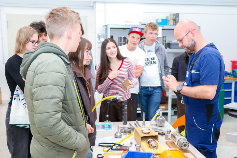Sieben Mädchen und Jungen stehen an einem technischen Prüfaufbau in einem Schulungsraum und betrachten verschiedene Bauteile und Messgeräte auf einem Arbeitstisch. Ein Mann in SLV-Kleidung demonstriert ein kleines Bauteil, im Hintergrund sind weitere technische Geräte und Arbeitsplätze zu sehen.