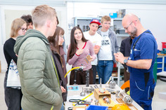 Sieben Mädchen und Jungen stehen an einem technischen Prüfaufbau in einem Schulungsraum und betrachten verschiedene Bauteile und Messgeräte auf einem Arbeitstisch. Ein Mann in SLV-Kleidung demonstriert ein kleines Bauteil, im Hintergrund sind weitere technische Geräte und Arbeitsplätze zu sehen.